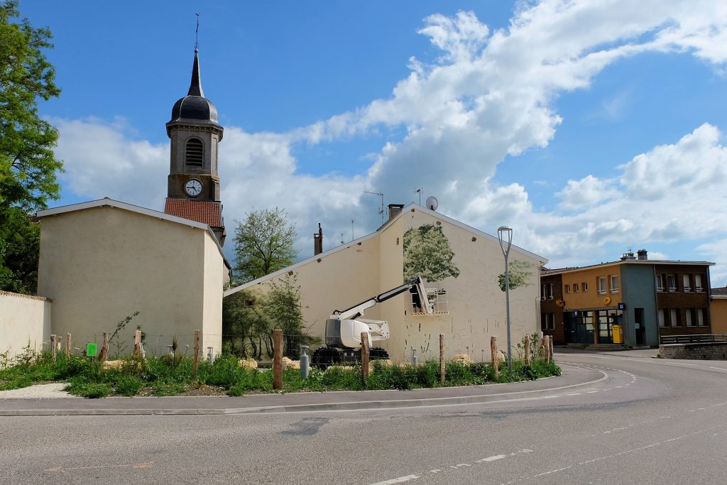 Vue depuis la rue de l’œuvre en cours de production. Une forêt et un feuillage peints commencent à apparaitre sur les murs du bâtiment.