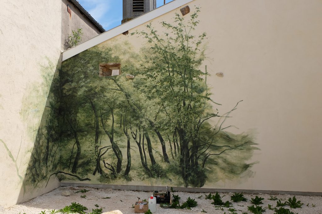 Vue rapprochée de l’œuvre en cours de production. Un pan de mur est peint avec un motif de forêt. Une forêt et un feuillage peints commencent à apparaitre sur les murs du bâtiment.