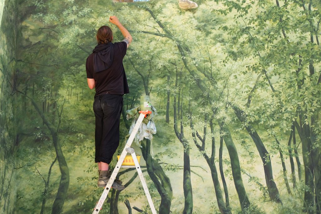Vue très rapprochée de l’œuvre en cours de production. L’artiste Noel Varoqui est de dos, en train de peindre la forêt.