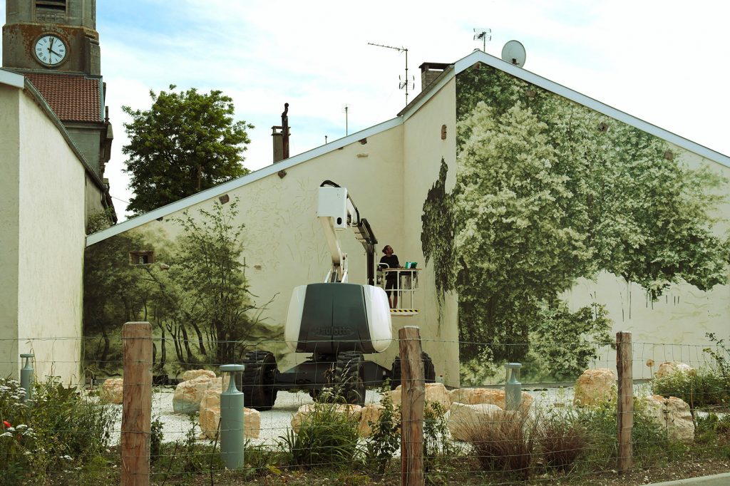 Vue d'ensemble de l’œuvre en cours de production. L’artiste Noel Varoqui est sur une nacelle devant le bâtiment devant un grand feuillage en cours de réalisation.