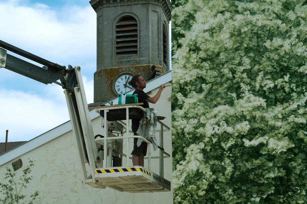 Vue rapprochée de Noel Varoqui, sur une nacelle, qui peint l’œuvre. Un morceau de mur est peint, le clocher de l’église apparait derrière lui.