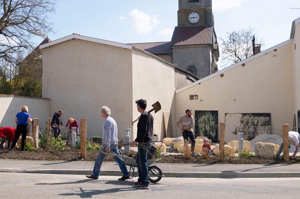 Vue d’ensemble du bâtiment avant qu’il soit peint. Plusieurs personnes préparent le terrain et jardinent devant les murs du bâtiment.