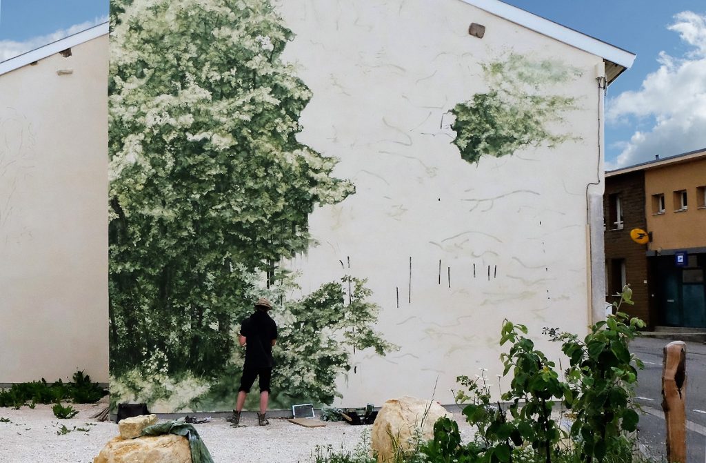 Vue de l’œuvre en cours de production, Noel Varoqui peint sur le mur, une forêt apparait sur la gauche du mur, des traits apparaissent sur le reste du mur.