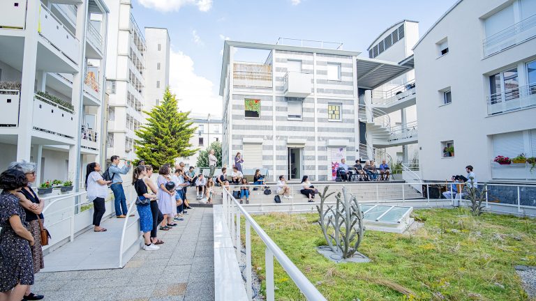 Vue de l’inauguration dans la cour de la résidence. Les sculptures sont disposées sur l’herbe dans l’espace central. Le public est autour