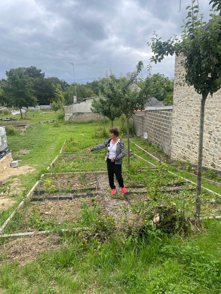 Nigel Bole, herboriste, se tenant au centre d'un jardin, entourée de plantes, sur la parcelle de L'Autre regard aux Prairies Saint-Martin.