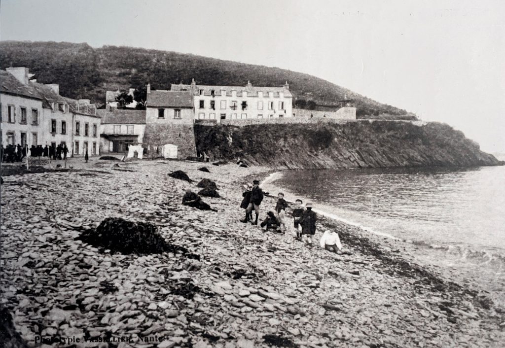 Image d'archive en noir et blanc du village de Maison Blanche vers 1900. Vue de la plage de galets où se trouve un groupe de personne et le village à gauche.