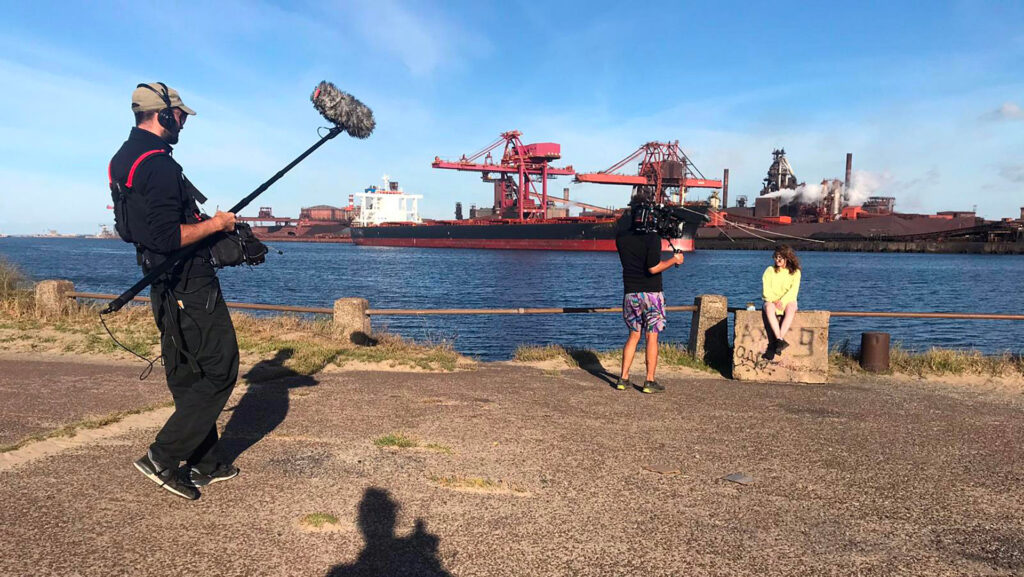 un caméraman et un perchiste filment et prennent le son lors du tournage. Une jeune fille assise sur un plot près de lot est face à eux. Dans l’arrière-plan, un cargo industriel est sur l’eau.