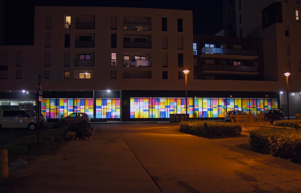 Vue de nuit du rideau coloré, déployé sur la façade vitrée. Des formes rectangulaires colorées sont imprimées sur ce tissu de 30m de long.