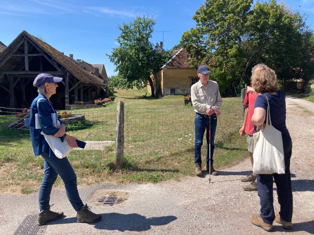 Un groupe de personnes, les commanditaires et l’une des médiatrices, se tient autour d'une clôture devant une grange, lors de la découverte du quartier des potiers.