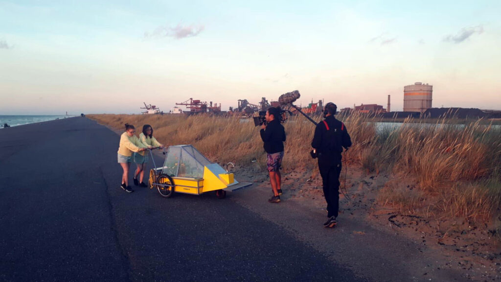 Deux jeunes filles en sweat-shirt jaunes poussent l’œuvre « Cataplasme » sur une route près de la mer, un caméraman et un perchman enregistrent le son et l’image.