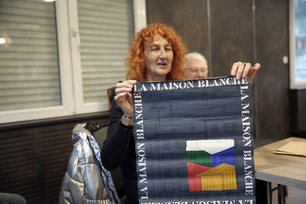 Une femme de la famille Gourmelon, tenant une affiche avec les mots « La Maison Blanche » et une illustration de maisons colorées, explique la vie quotidienne après-guerre.