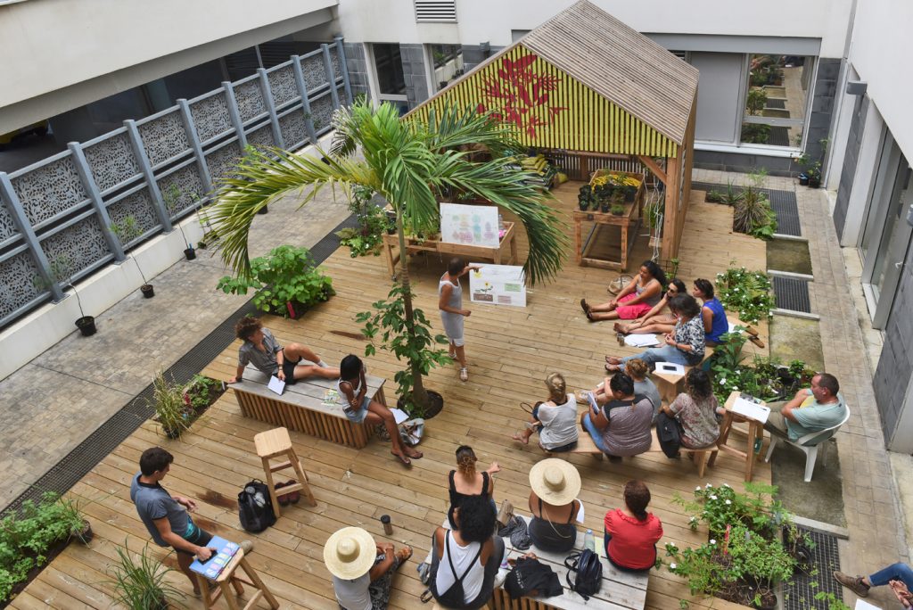 Dans la cour du CHU Saint Pierre, un groupe de personnes écoute un présentation dans le "Jardin la Nurserie". L'installation tout en vois est composée d'une terrasse centrale avec une structure avec une bâtisse ouverte et diverses plantes et mobiliers.