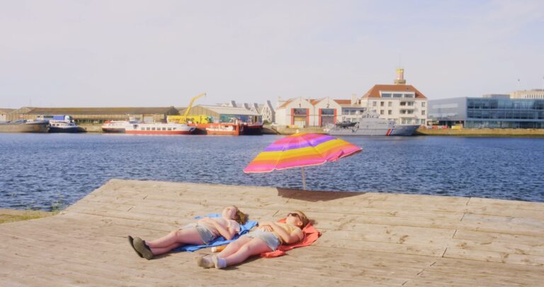 Deux jeunes filles sont allongées au soleil sur une terrasse en bois près de l’eau. On aperçoit derrière, après la mer, des bateaux industriels et un bateau de la marine, ainsi que des bâtiments.