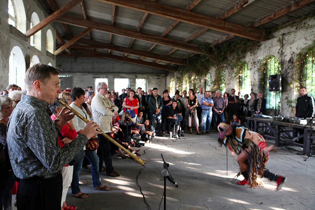 Dans un bâtiment, une performance chorégraphique d'une femme habillée en costume de chamane accompagnée de musiciens (un flutiste en premier plan), devant un public.