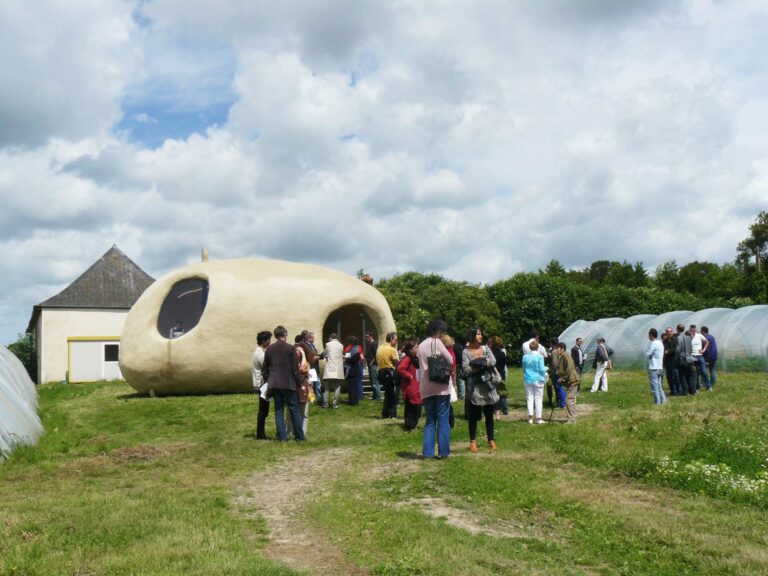 Des gens sont rassemblés dans les Jardins du Breil. Ils discutent dehors, entre les serres et la structure de Nicolas Floc'h.
