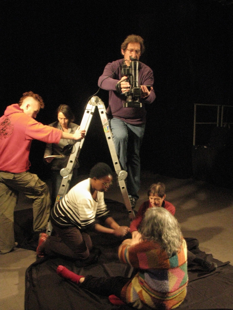 Trois femmes assissent en cercle sur le sol. Leurs mains sont positionnées au centre de ce cercle. Derrière, sur un escabeau, un homme filme le mouvement de leurs mains.