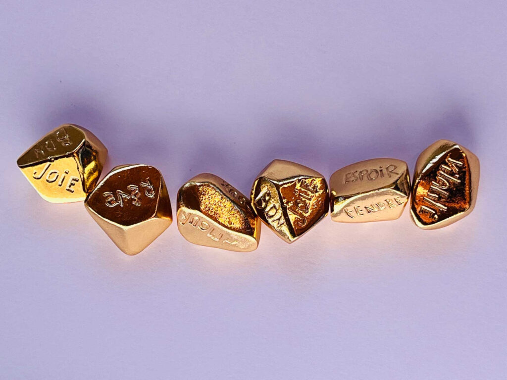 Osselets dorés sur lesquels sont notés des mots du champ lexical de la douceur et du bonheur (joie, rêve, bon, espoir, tendre...)